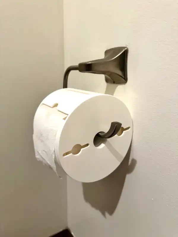 White Toilet Paper Protector covering a roll on a wall-mounted spring-arm holder, paper coming out of the slot